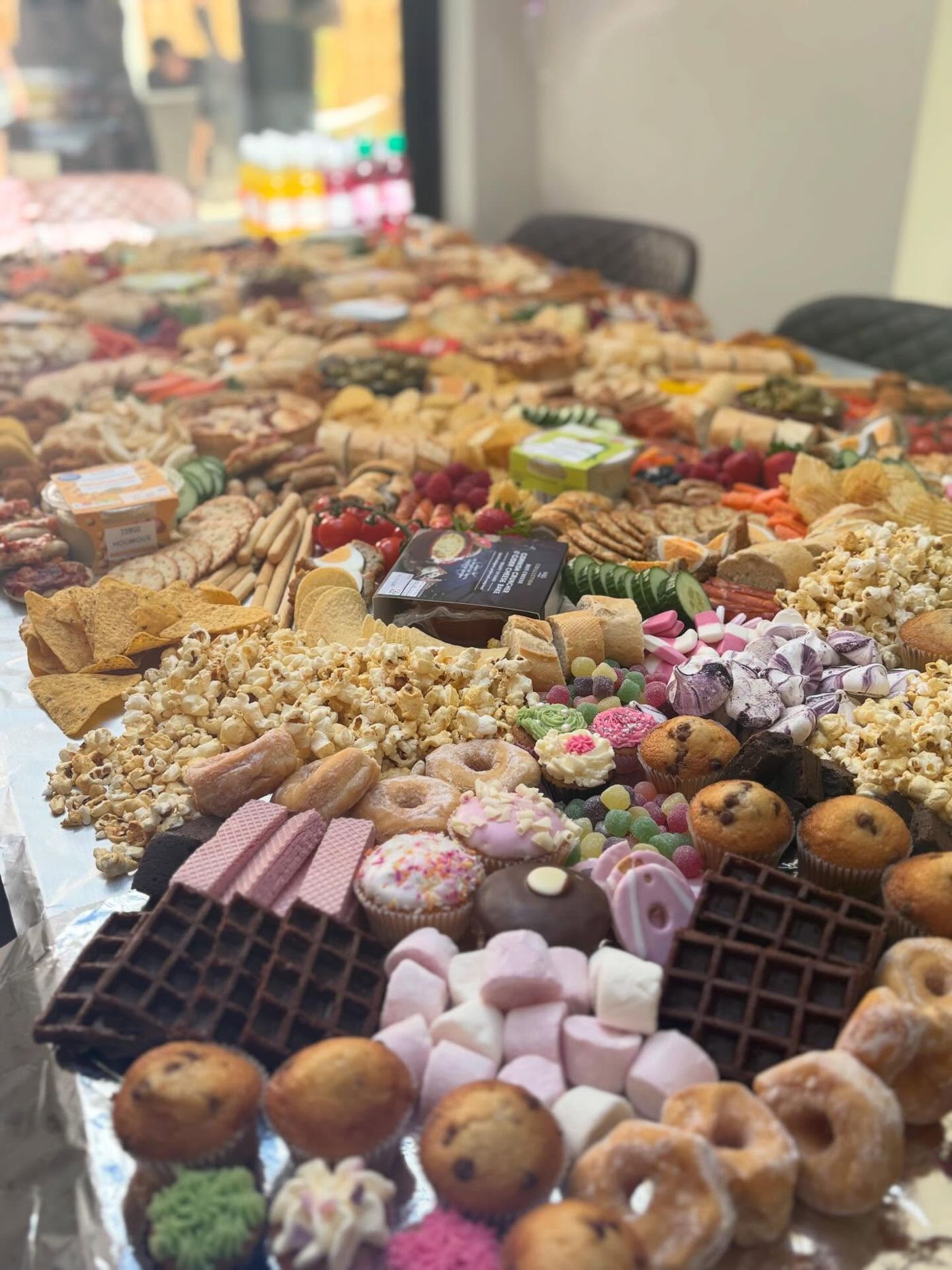 A very long party table of doughnuts, muffins, marshmallows and popcorn, with savoury food further back