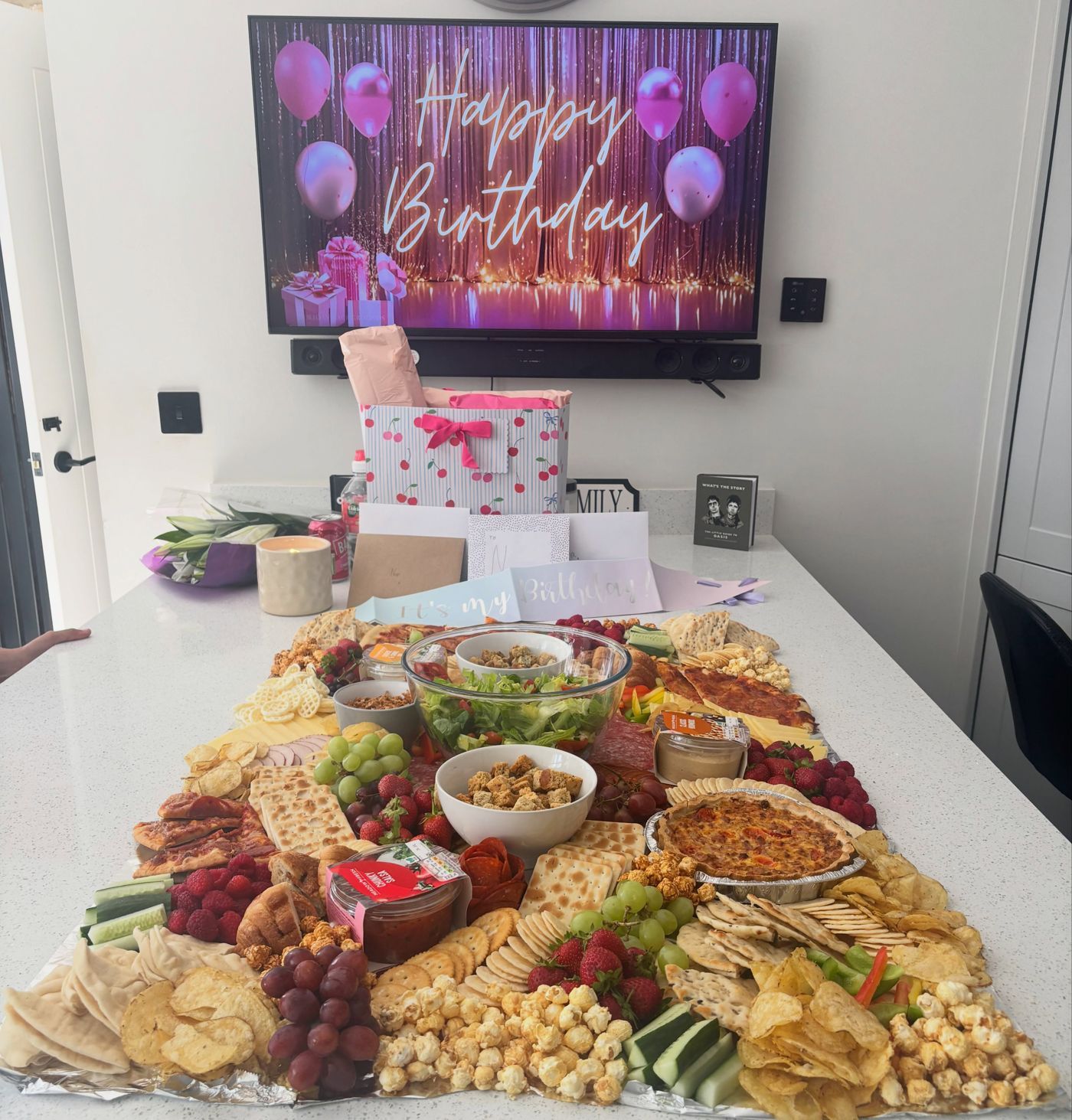 A grazing table on a white kitchen island in front of a television showing Happy Birthday