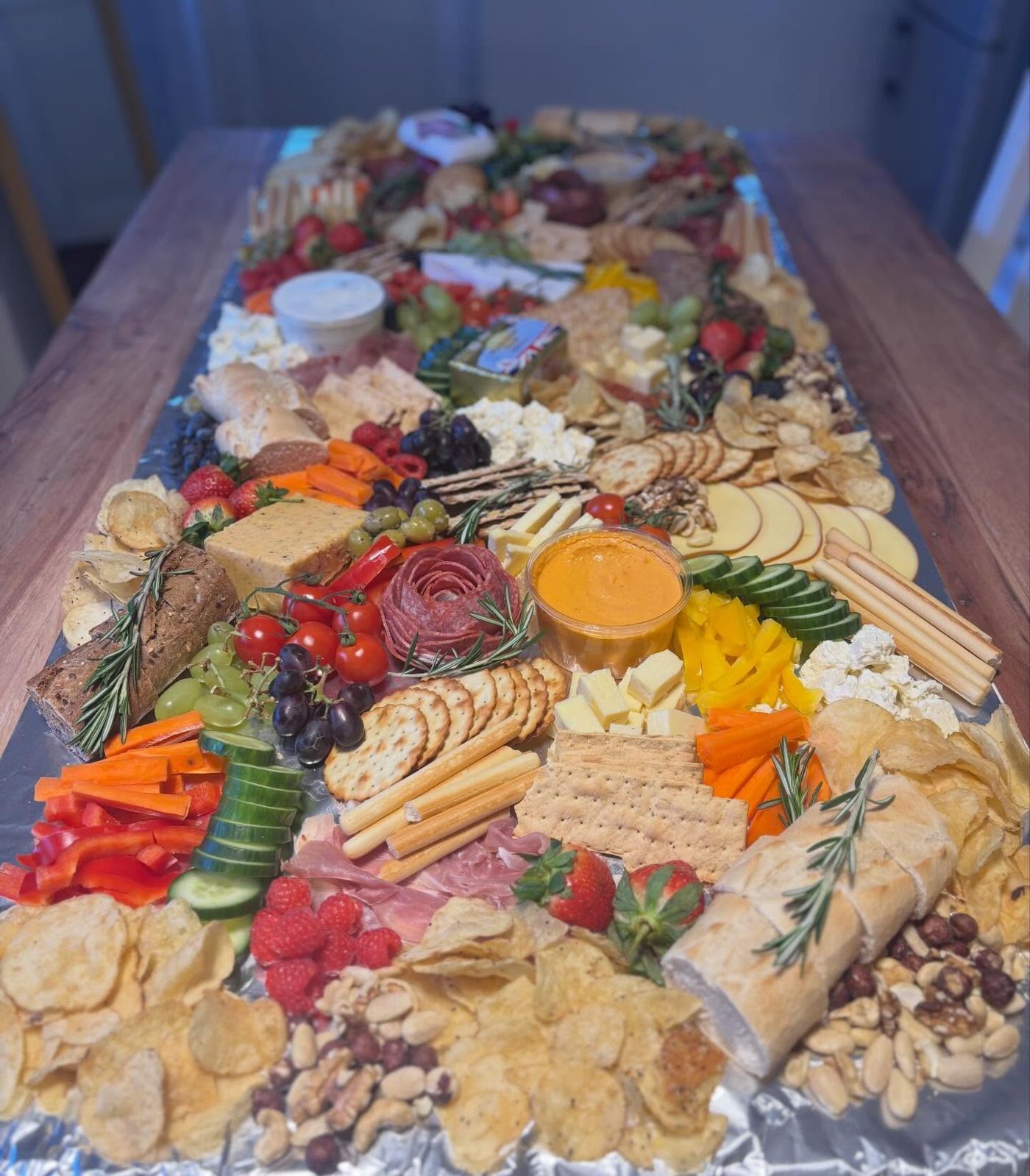 A grazing table running the length of a wooden dining table: cheeses, breads, crackers, berries, peppers and a salami rose, with sprigs of rosemary